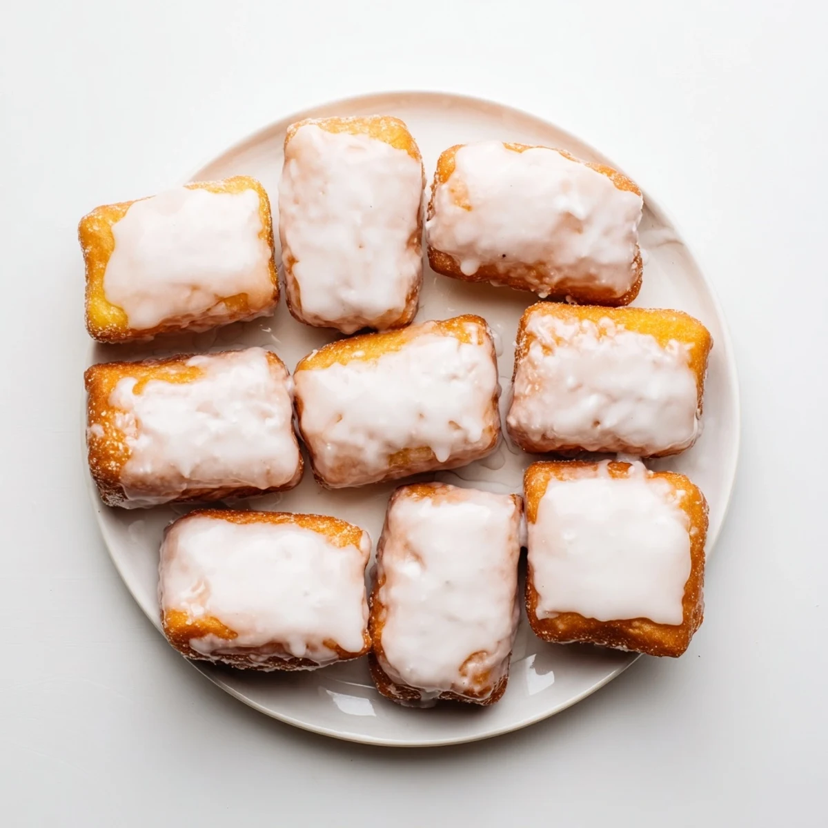 Golden glazed buttermilk beignet squares stacked on a white serving plate with vanilla glaze dripping down the sides