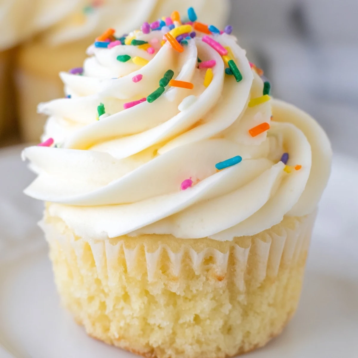 Golden vanilla cupcakes topped with creamy swirls of vanilla buttercream frosting on a white plate
